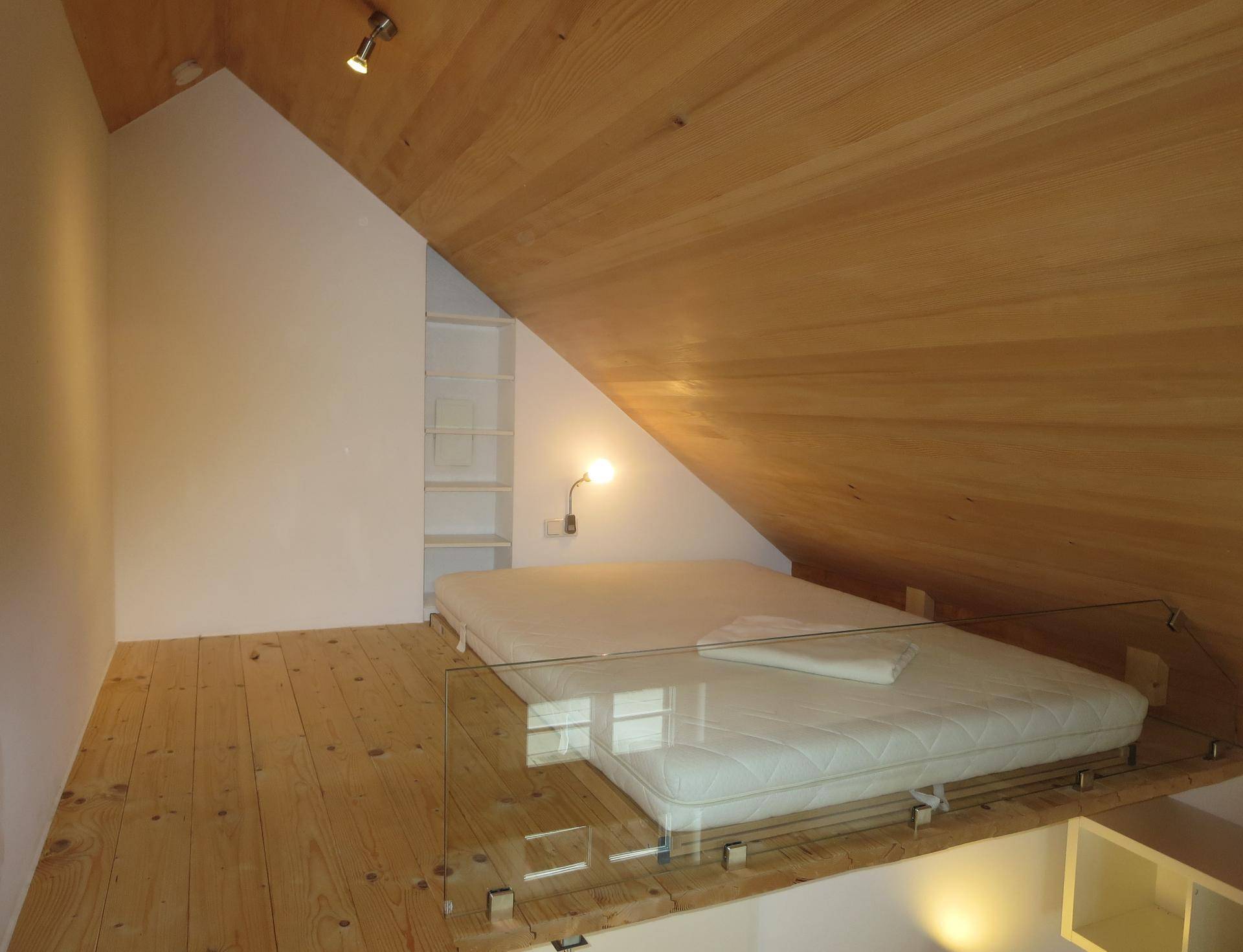 Gemütlicher Schlafbereich im Dachgeschoss mit Holzwänden, schrägen Decken und Glasgeländer.