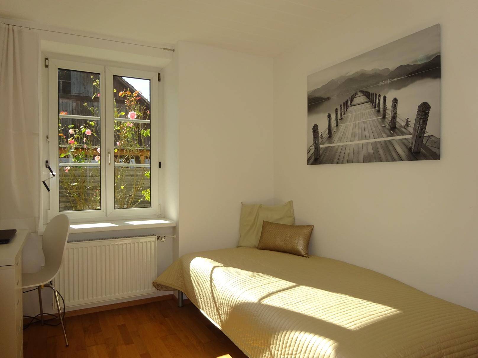 Helles Schlafzimmer mit Einzelbett, Schreibtisch und Blick ins Grüne durch das Fenster.