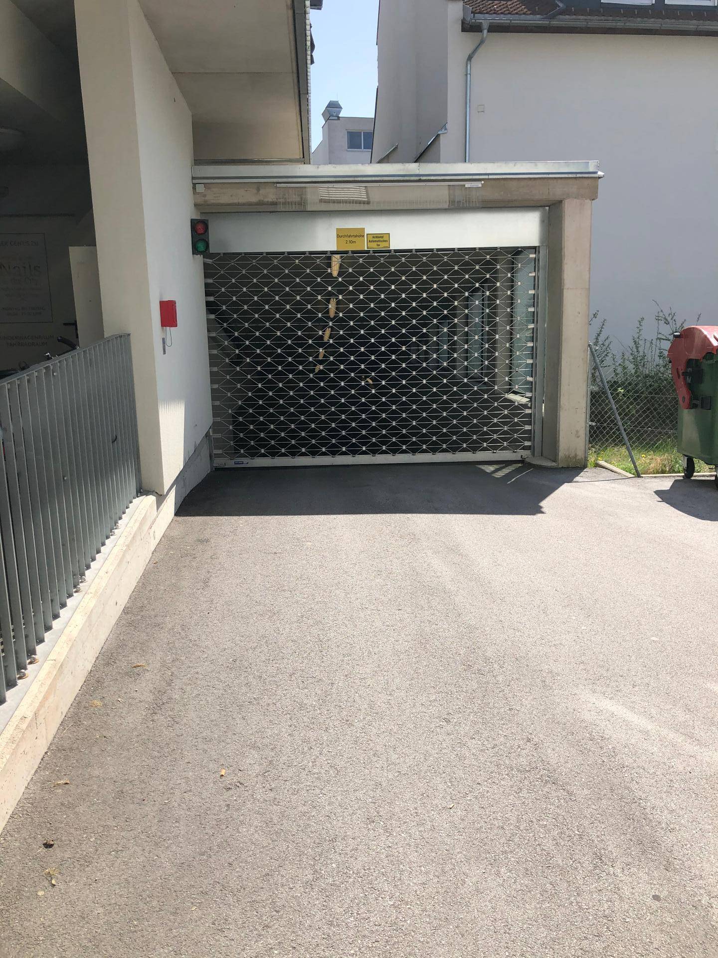 Einfahrt zu einer Tiefgarage mit geschlossenem Rolltor an einem sonnigen Tag.