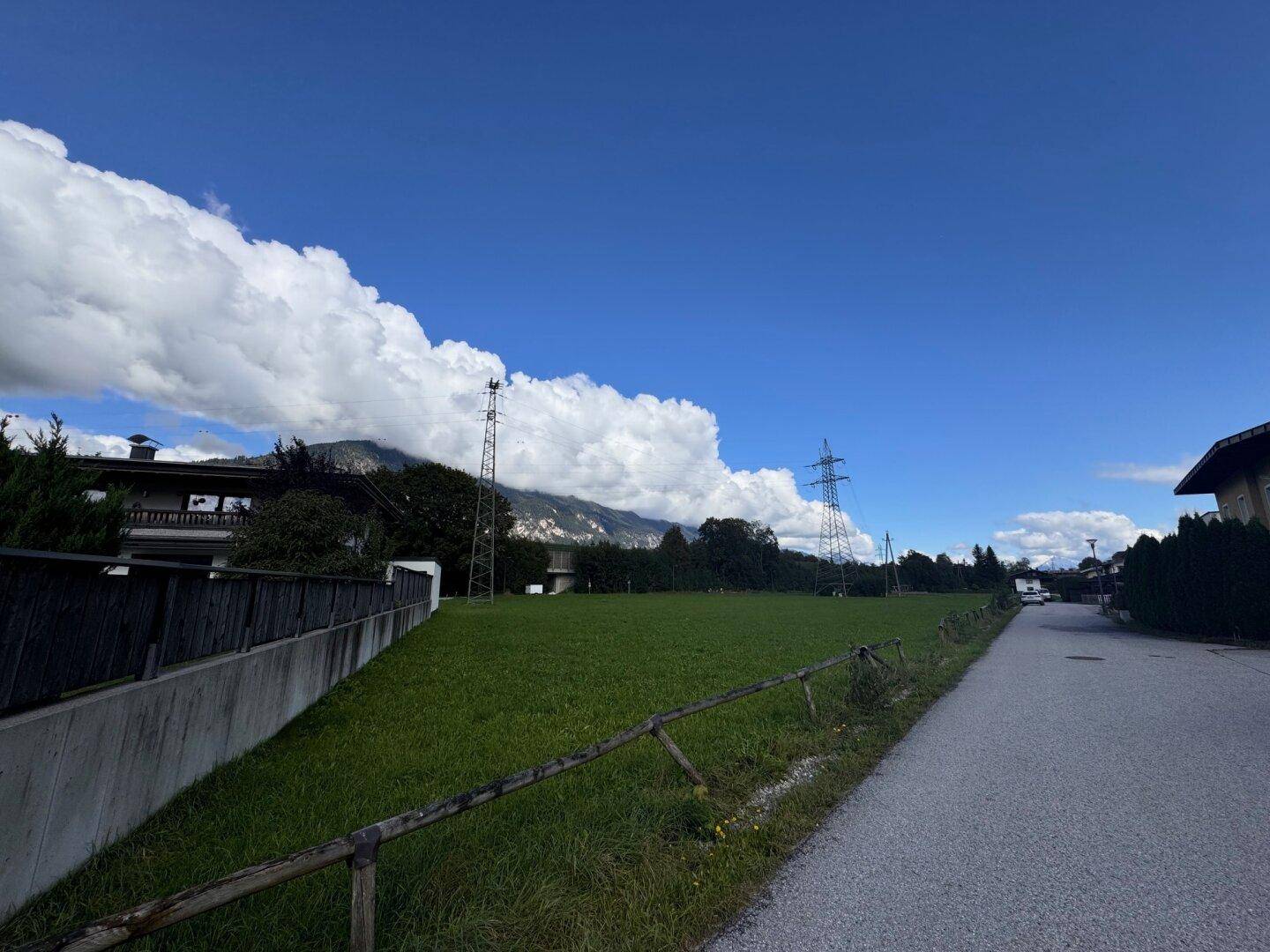 Weitläufige grüne Fläche neben einer Straße mit einem Wohngebäude und Bergpanorama im Hintergrund.