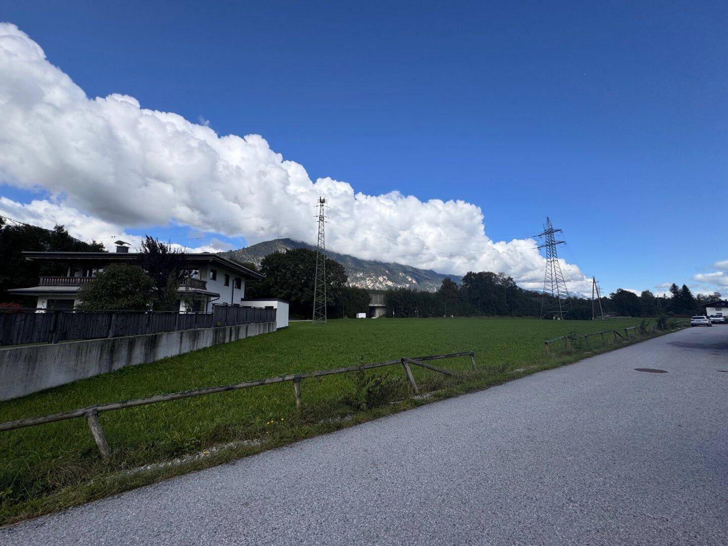 Grüne Landschaft neben einer Straße mit einem Wohnhaus und Bergen unter einem weiten blauen Himmel.