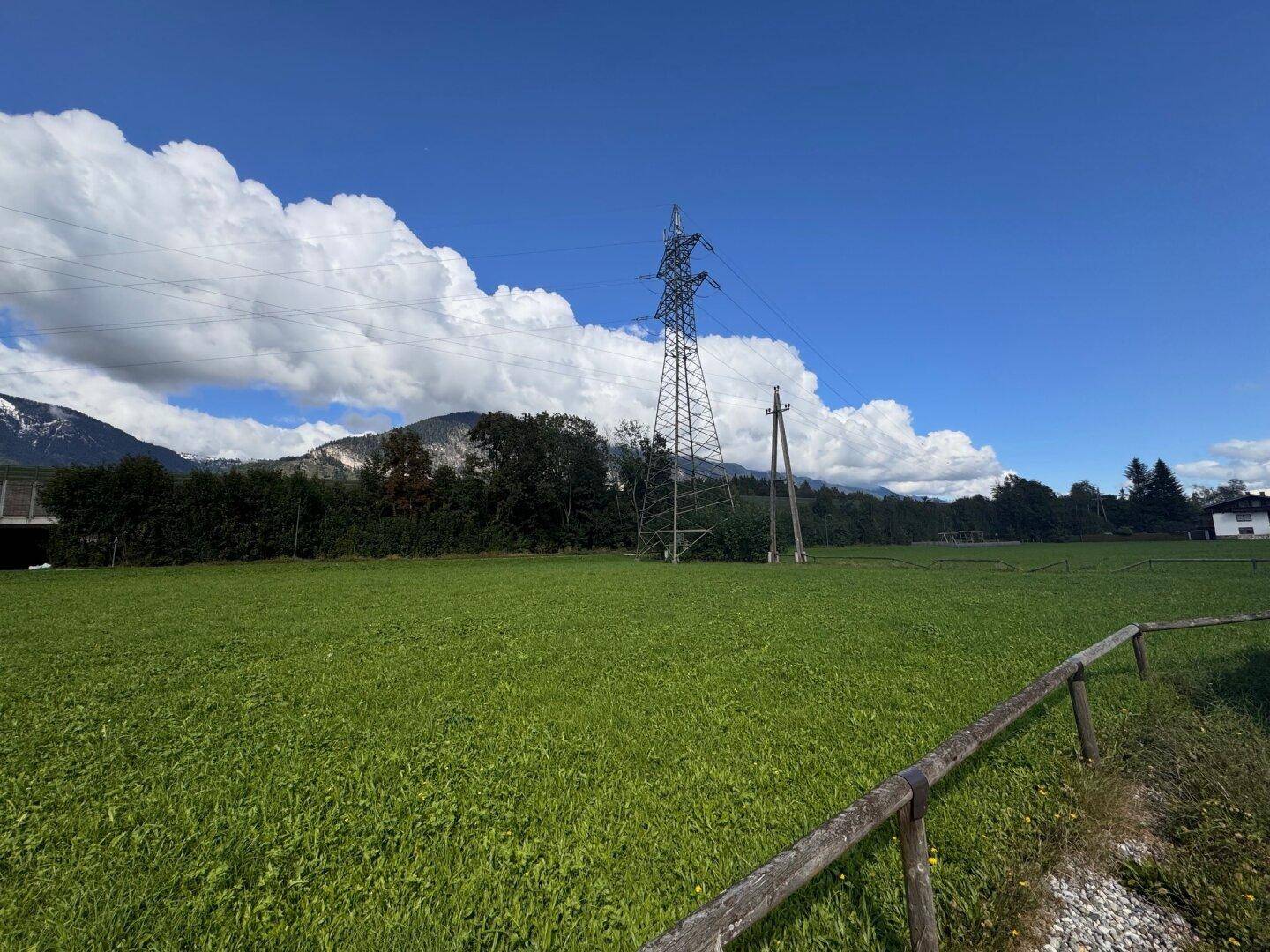 Grüne Wiese mit einem großen Strommast im Vordergrund und einer beeindruckenden Bergkulisse dahinter.