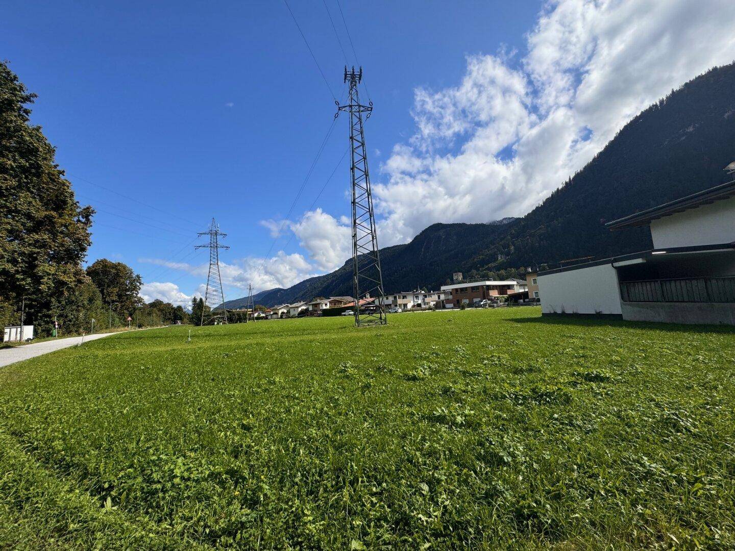Weite grüne Fläche mit einem Strommast, umgeben von Wohnhäusern und einer majestätischen Bergkette.