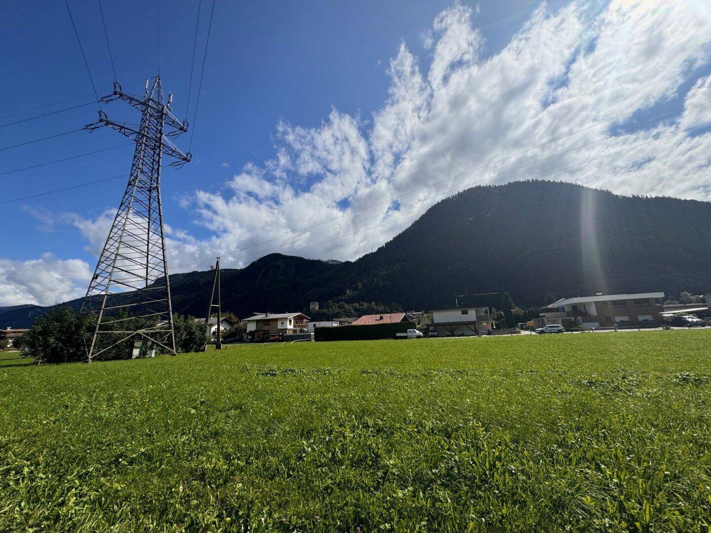 Grüne Wiese mit einem hohen Strommast und Häusern im Hintergrund, umgeben von bewaldeten Bergen.