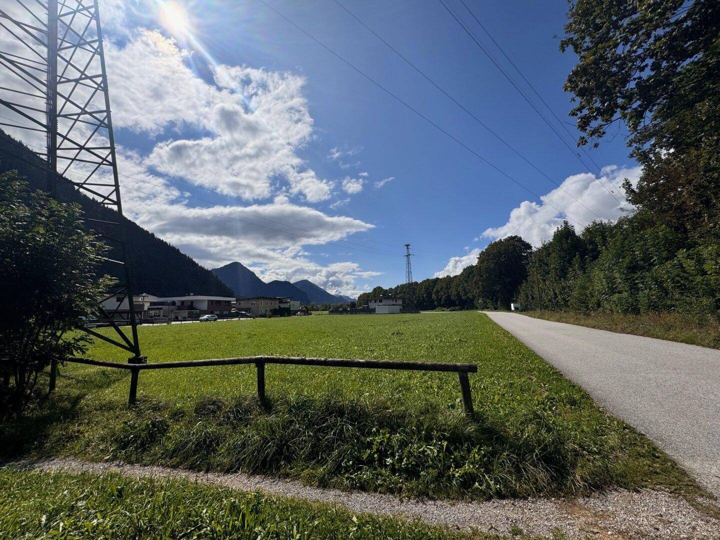 Sonniger Blick über eine grüne Wiese mit einem Strommast und einer Straße, umgeben von Bergen.