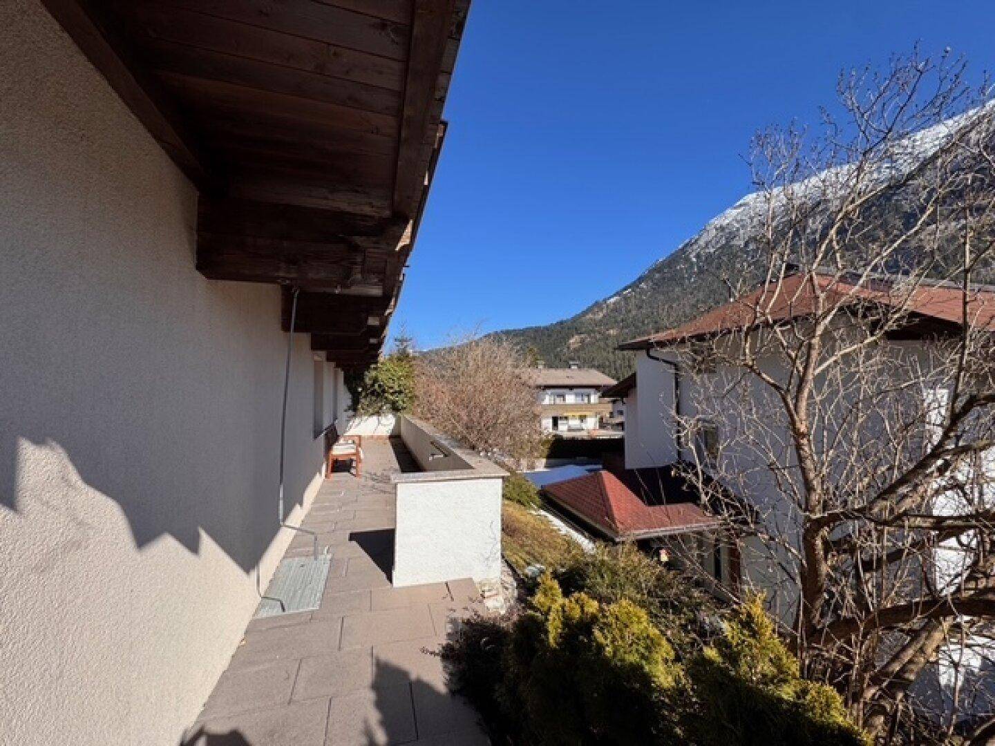 Langer Balkon mit Fliesenboden und Ausblick auf die umliegenden Häuser und Berge.