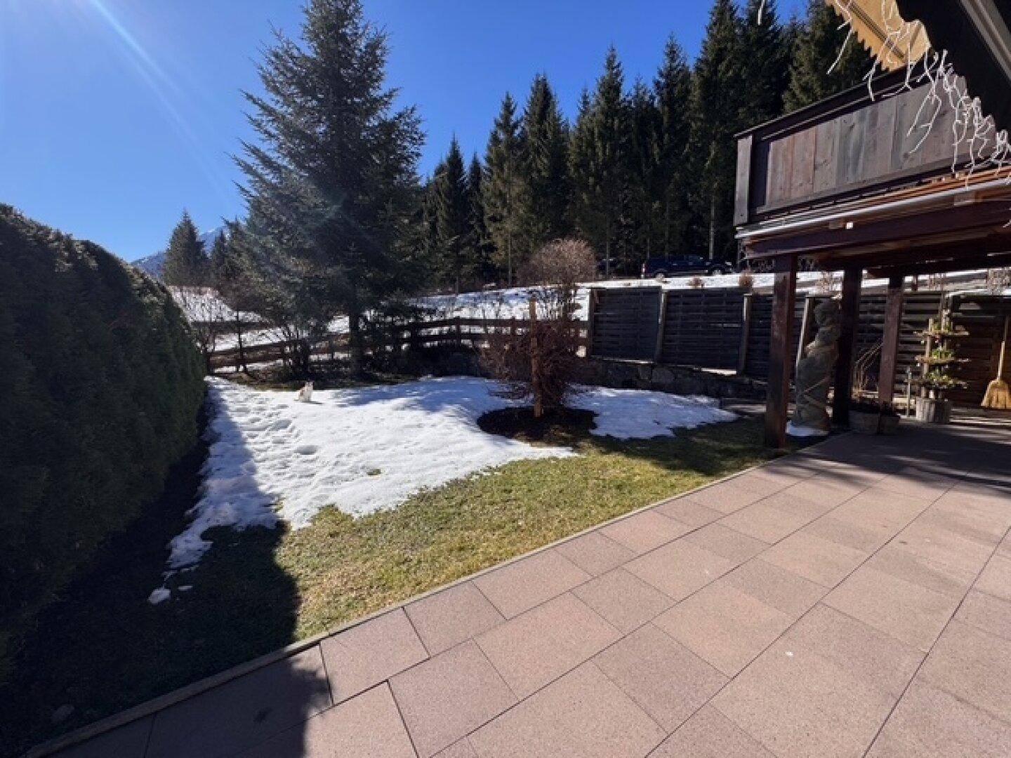 Gartenbereich mit Rasen, Schnee und gepflastertem Weg, umgeben von Bäumen und Hecken.