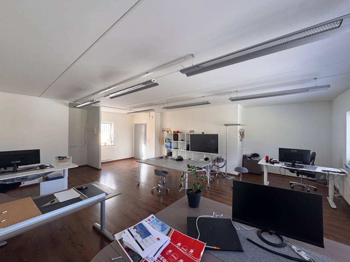 Offener Bürobereich mit funktionalen Arbeitsplätzen, Regalsystemen und Zugang zu einem weiteren Raum.