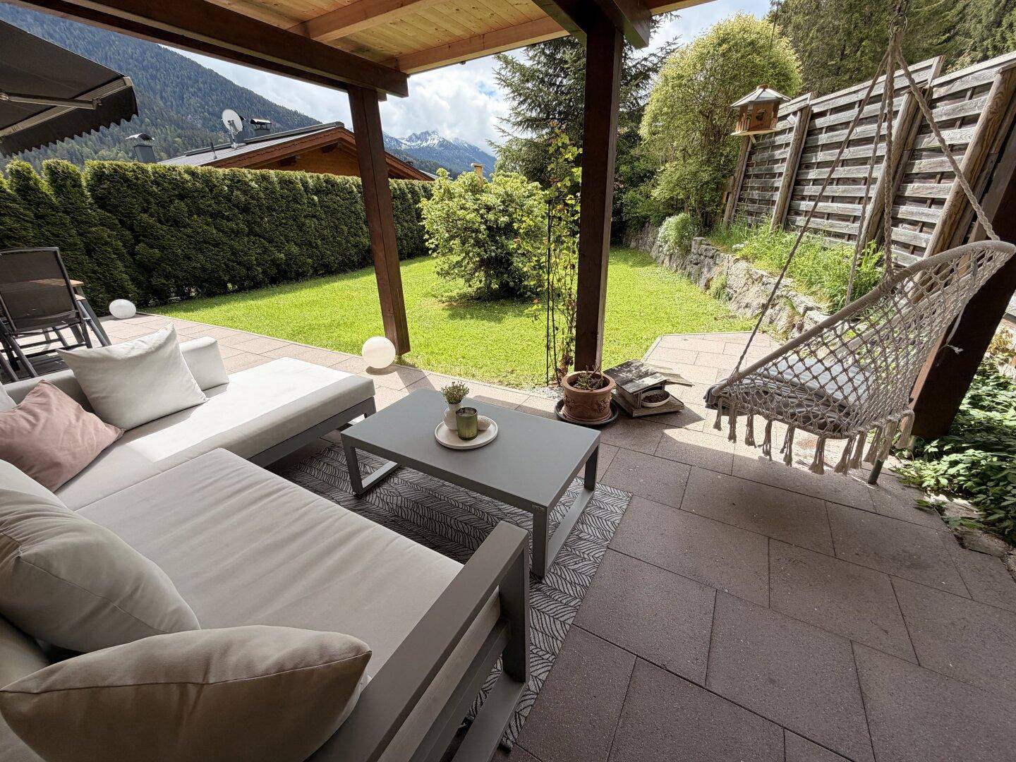 Überdachte Terrasse mit bequemen Lounge-Möbeln, Hängesessel und Blick auf den Garten und die Berge.