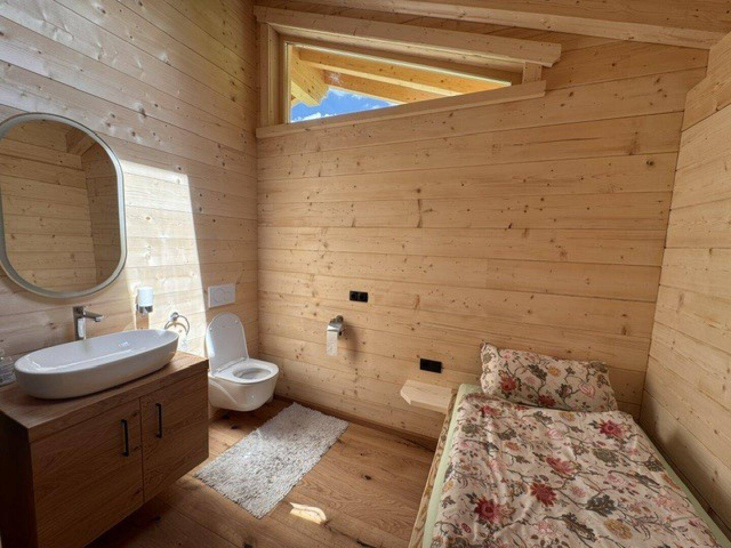 Gemütliches Schlafzimmer mit Holzwänden, Einzelbett, Waschbecken und wandhängendem WC.