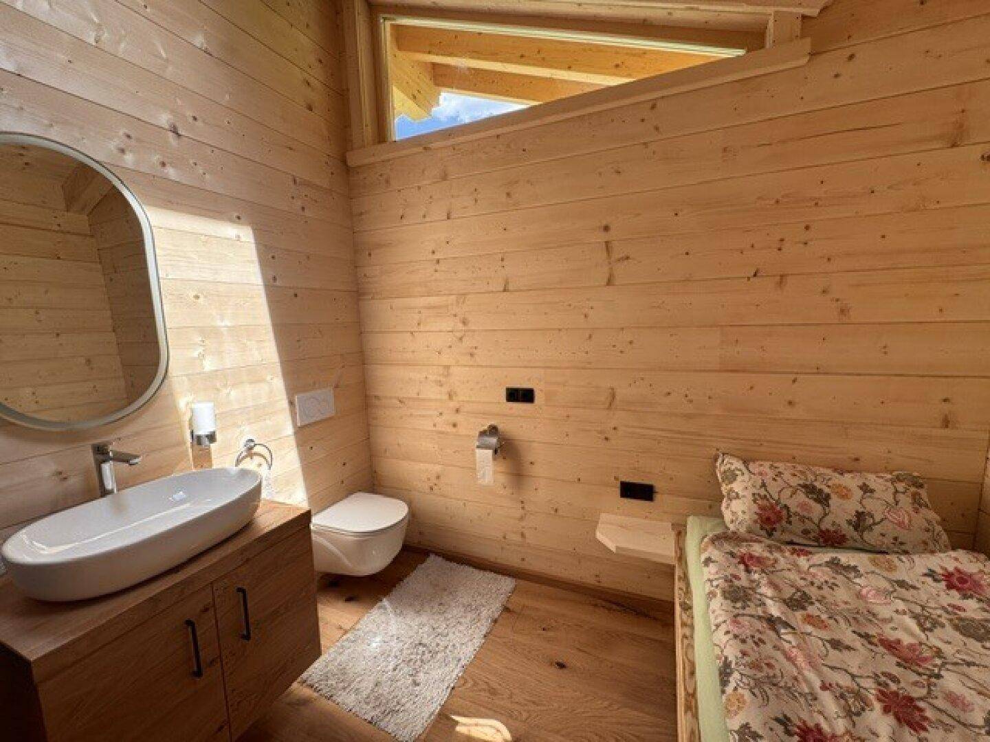 Lichtdurchflutetes Schlafzimmer mit Holzwänden, Einzelbett, Waschbecken und WC, ideal für Gäste.