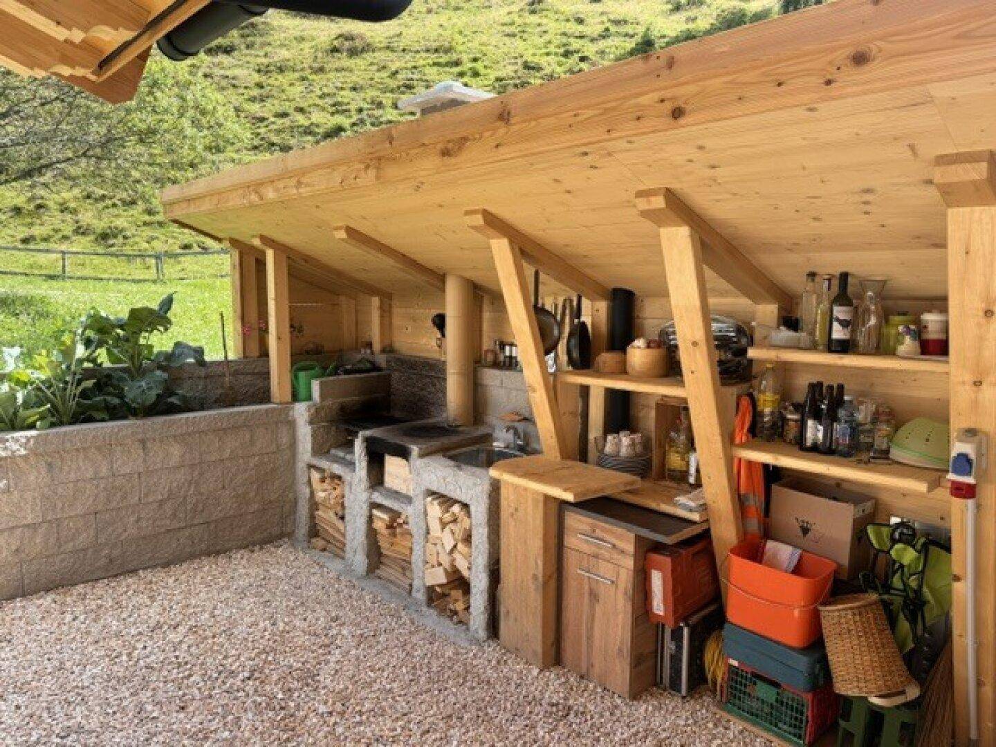 Überdachter Außenbereich mit Grillplatz, Holzregalen und Blick auf die grüne Berglandschaft.