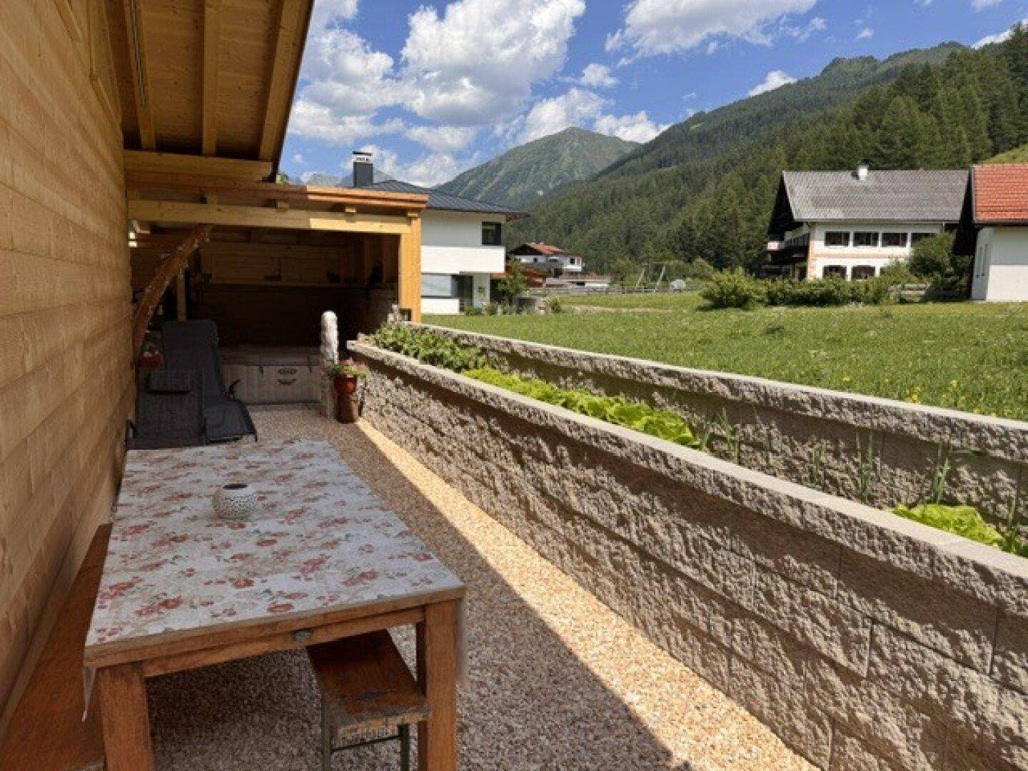 Sonniger Außenbereich mit Holztisch, Bänken und bepflanzten Hochbeeten vor Bergkulisse.