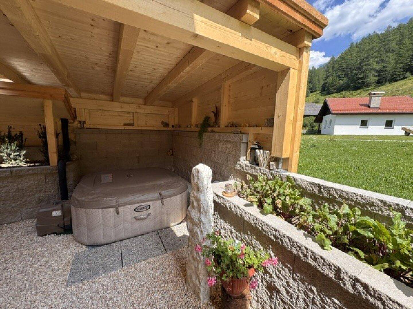 Überdachter Terrassenbereich mit aufblasbarem Whirlpool und bepflanzten Hochbeeten in alpiner Umgebung.