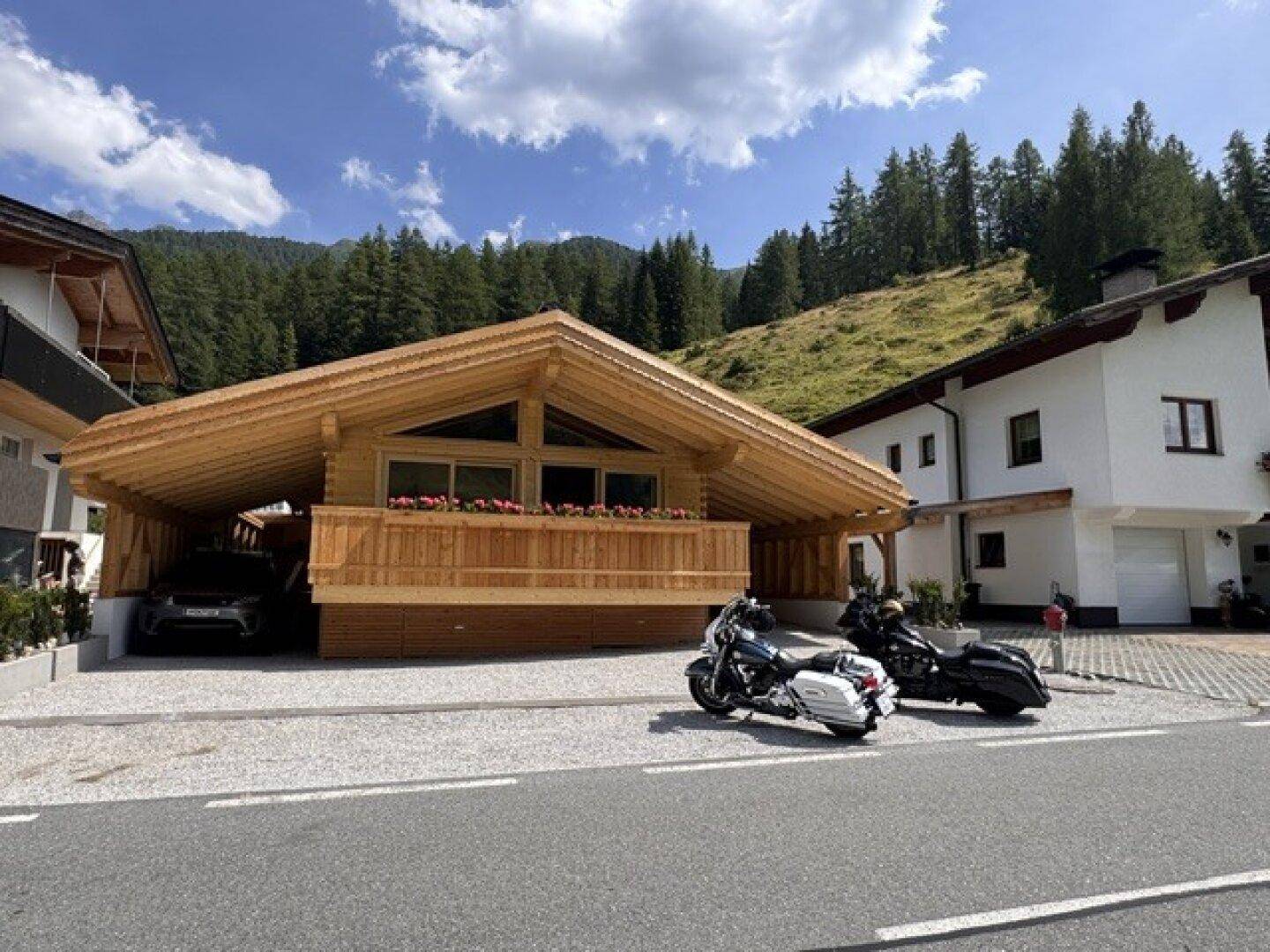 Modernes Holzchalet mit Carport und Balkon, eingebettet in eine Berglandschaft neben einer Straße.