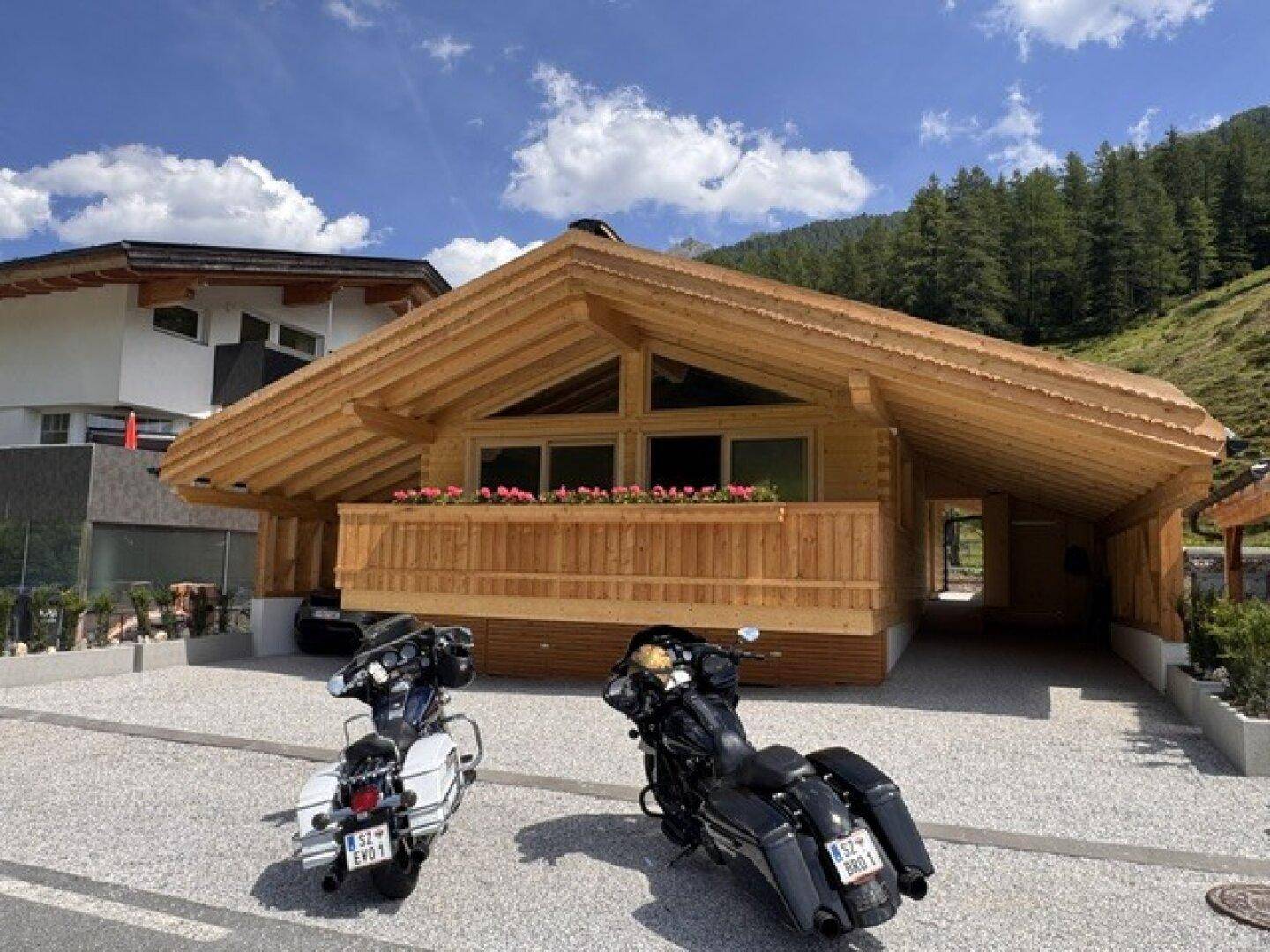 Neues Holzchalet mit Satteldach und Balkon, umgeben von grünen Hügeln und zwei Motorrädern.