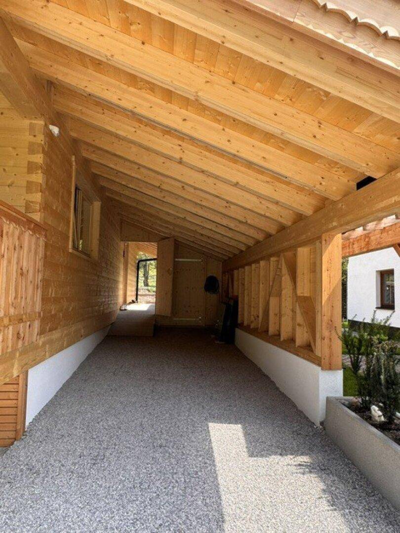 Heller Carport mit Holzbalken und Kiesboden, bietet geschützten Zugang zum Haus.