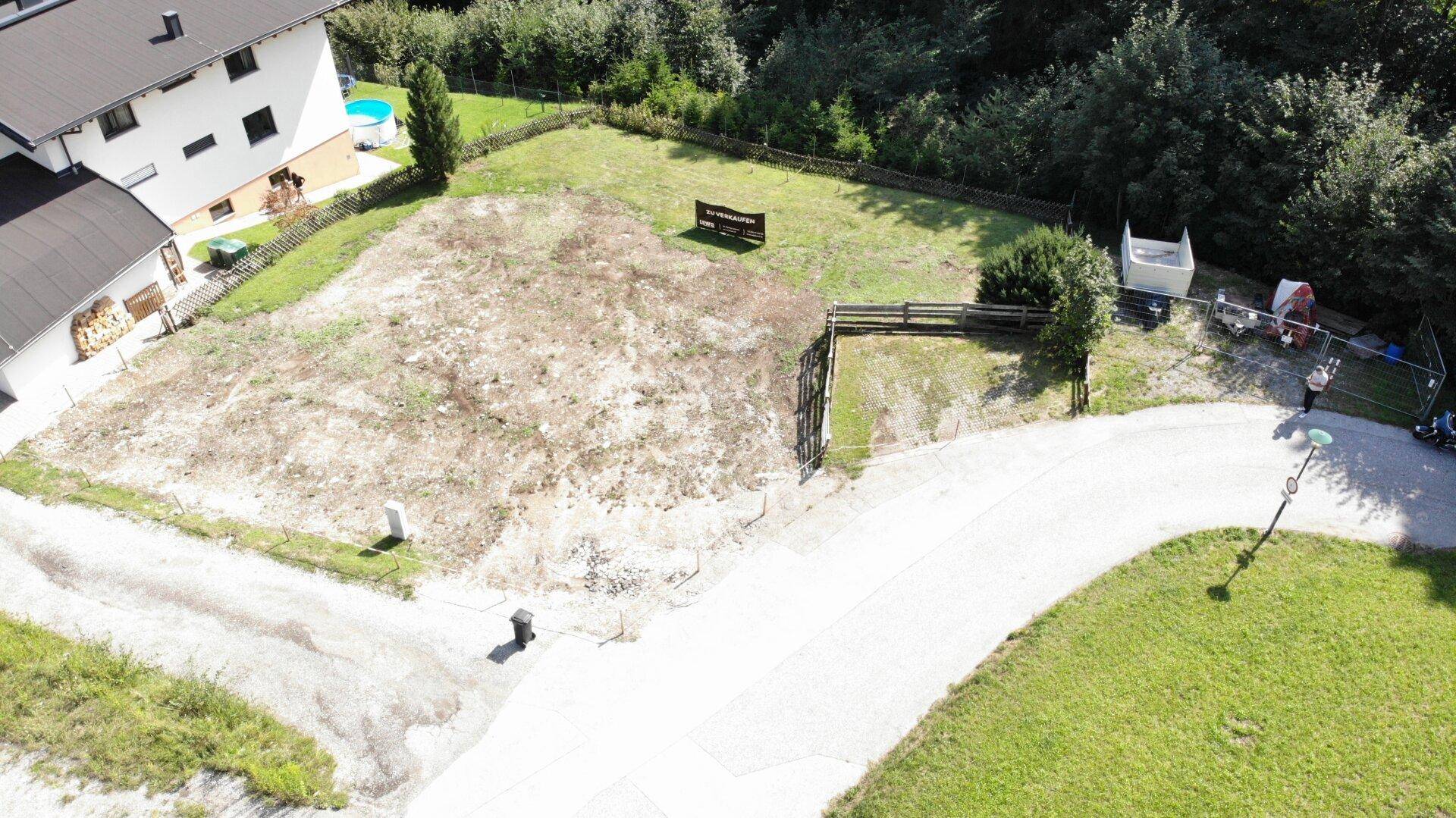 Luftaufnahme eines Baugrundstücks mit angrenzendem Haus und Pool, umgeben von viel Grün.