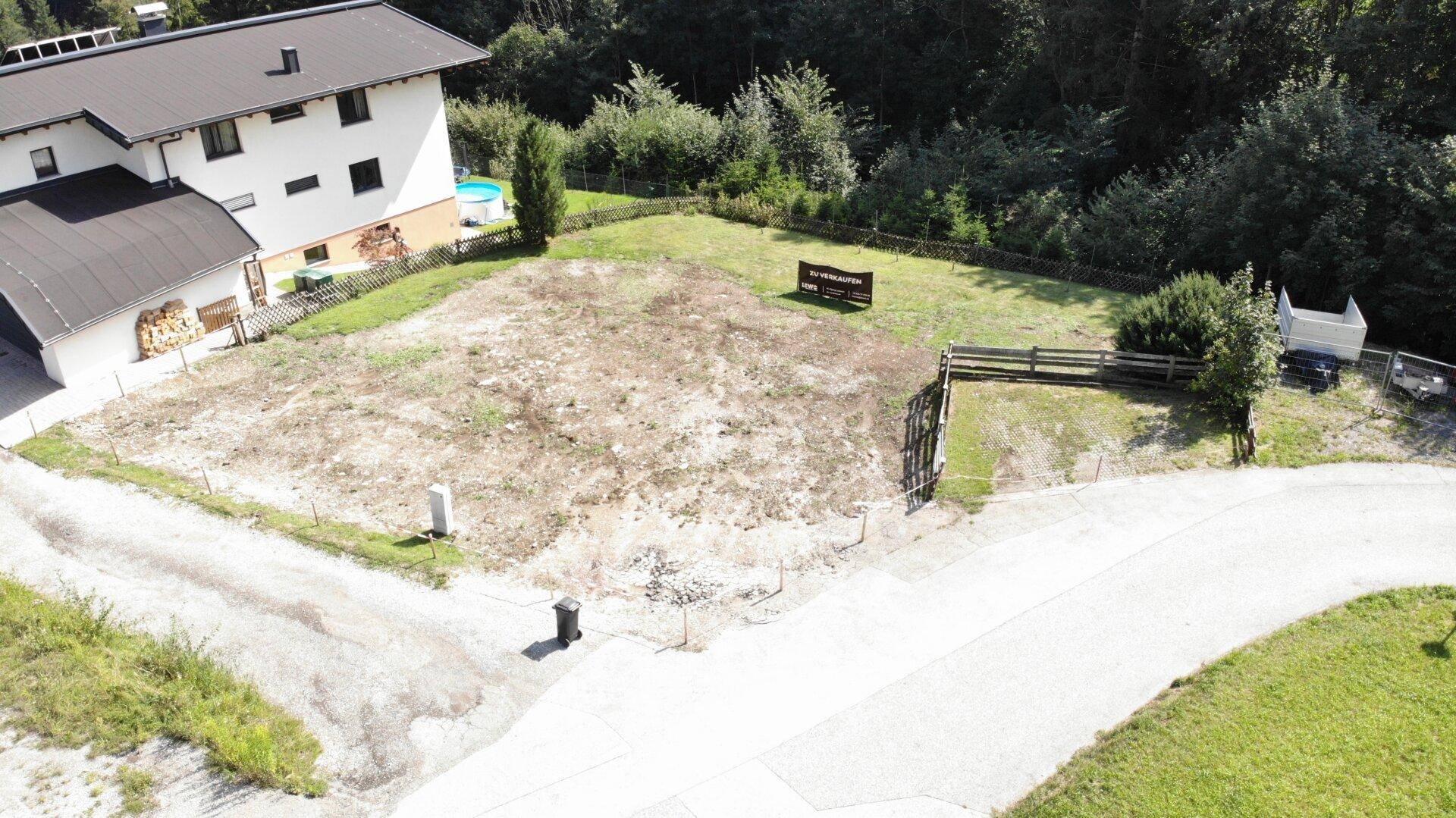 Überblick über ein Baugrundstück neben einem bestehenden Wohnhaus mit Holzstapel und Pool im Hintergrund.