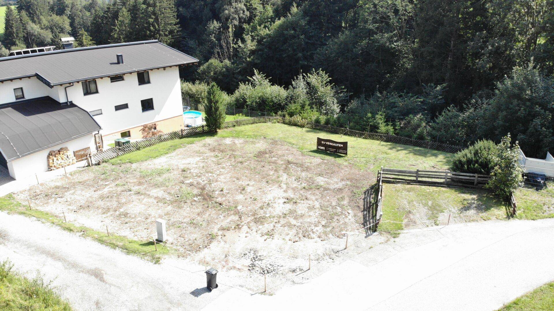 Überblick über ein Baugrundstück neben einem bestehenden Wohnhaus mit Holzstapel und Pool im Hintergrund.