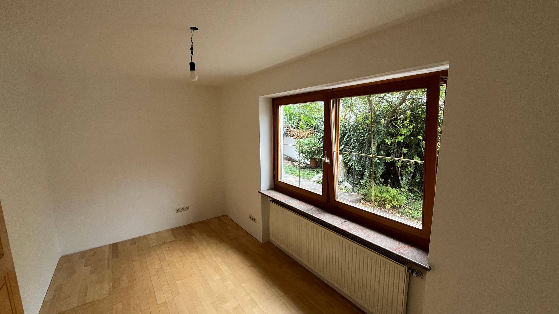 Helles Zimmer mit Parkettboden, großem Fenster und Blick in den grünen Garten.