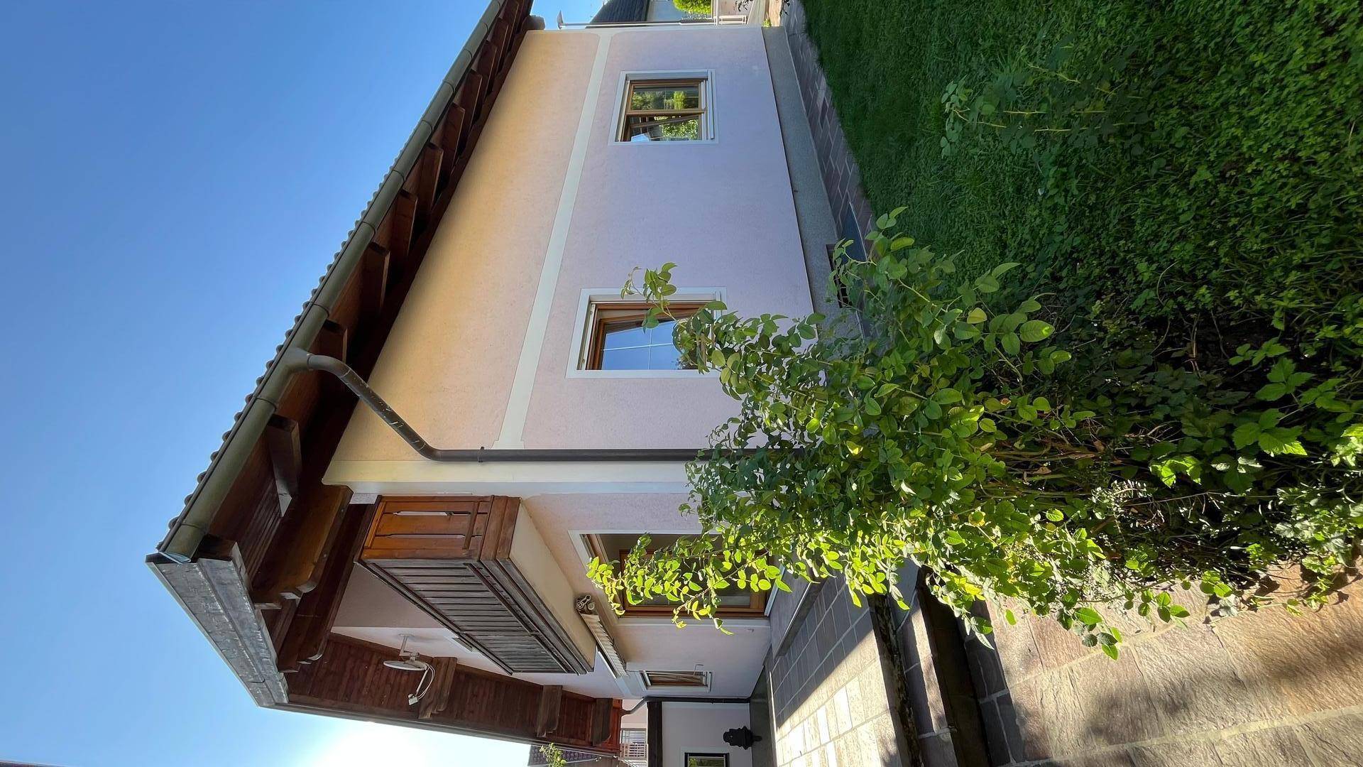 Seitenansicht eines gepflegten Hauses mit hellrosa Fassade, Holzbalkon und grünem Gartenbereich unter blauem Himmel.