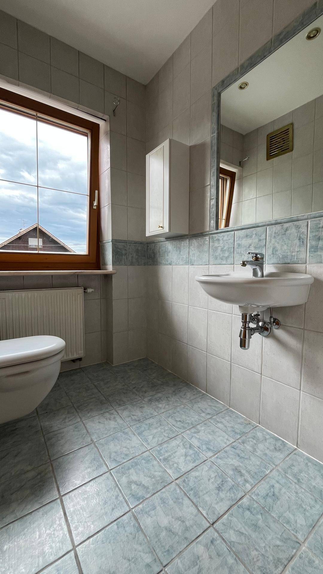 Kleines Badezimmer mit WC, Waschbecken, Fenster und hellblauen Fliesen.