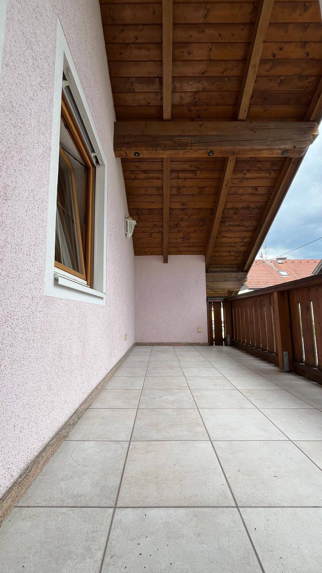 Langer Balkon mit Fliesenboden und Holzbalken unter einem bewölkten Himmel.