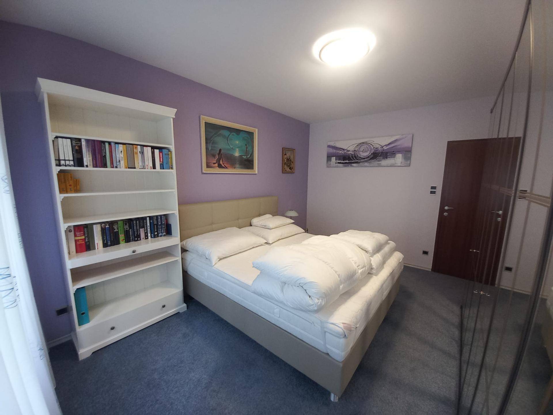 Geräumiges Schlafzimmer mit lila Wänden, einem Doppelbett und einem weißen Bücherregal für zusätzlichen Stauraum.