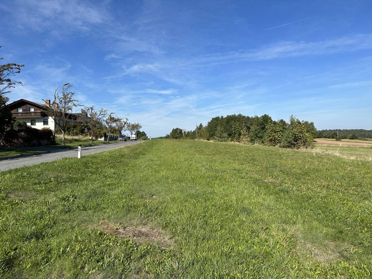 Grüne Wiese neben einer Landstraße mit einem Haus in der Ferne und bewaldeten Hügeln am Horizont.