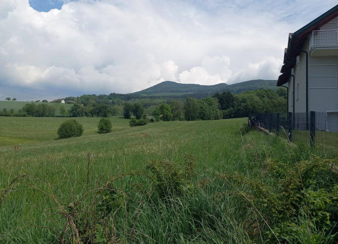 Modernes Gebäude mit Balkonen und weitem Blick über grüne Felder und bewaldete Berge unter bewölktem Himmel.