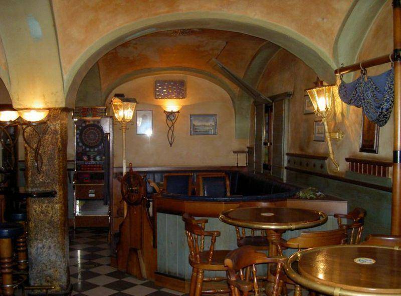 Gemütlicher Barbereich mit Holztischen, Stühlen und dekorativen Wandleuchten unter einem Gewölbedach und Fliesenboden.