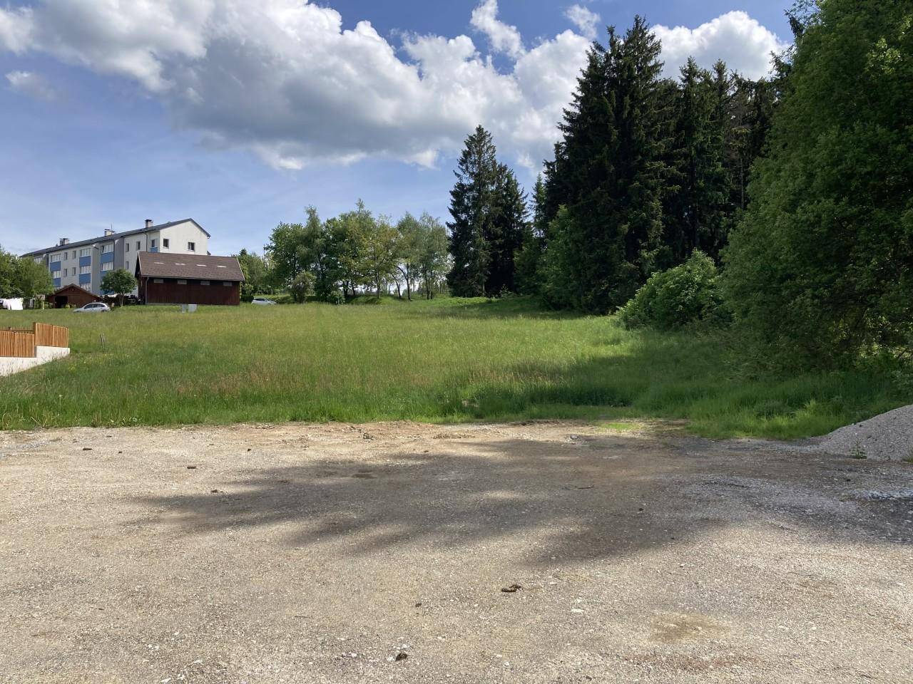 Offenes Grundstück mit Grasfläche, Schotter im Vordergrund und Bäumen sowie einem Gebäude in der Ferne.