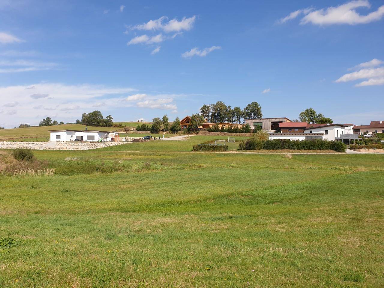 Panoramablick über eine grüne Wiesenlandschaft mit verstreuten modernen Häusern und bewaldeten Hügeln im Hintergrund.