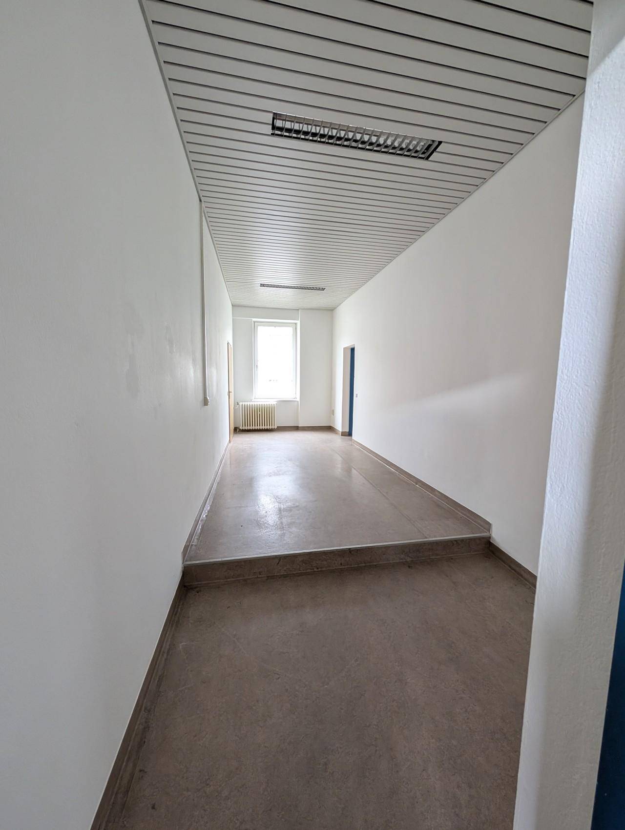 Langer, heller Flur mit Linoleumboden, weißen Wänden und einem Fenster am Ende.