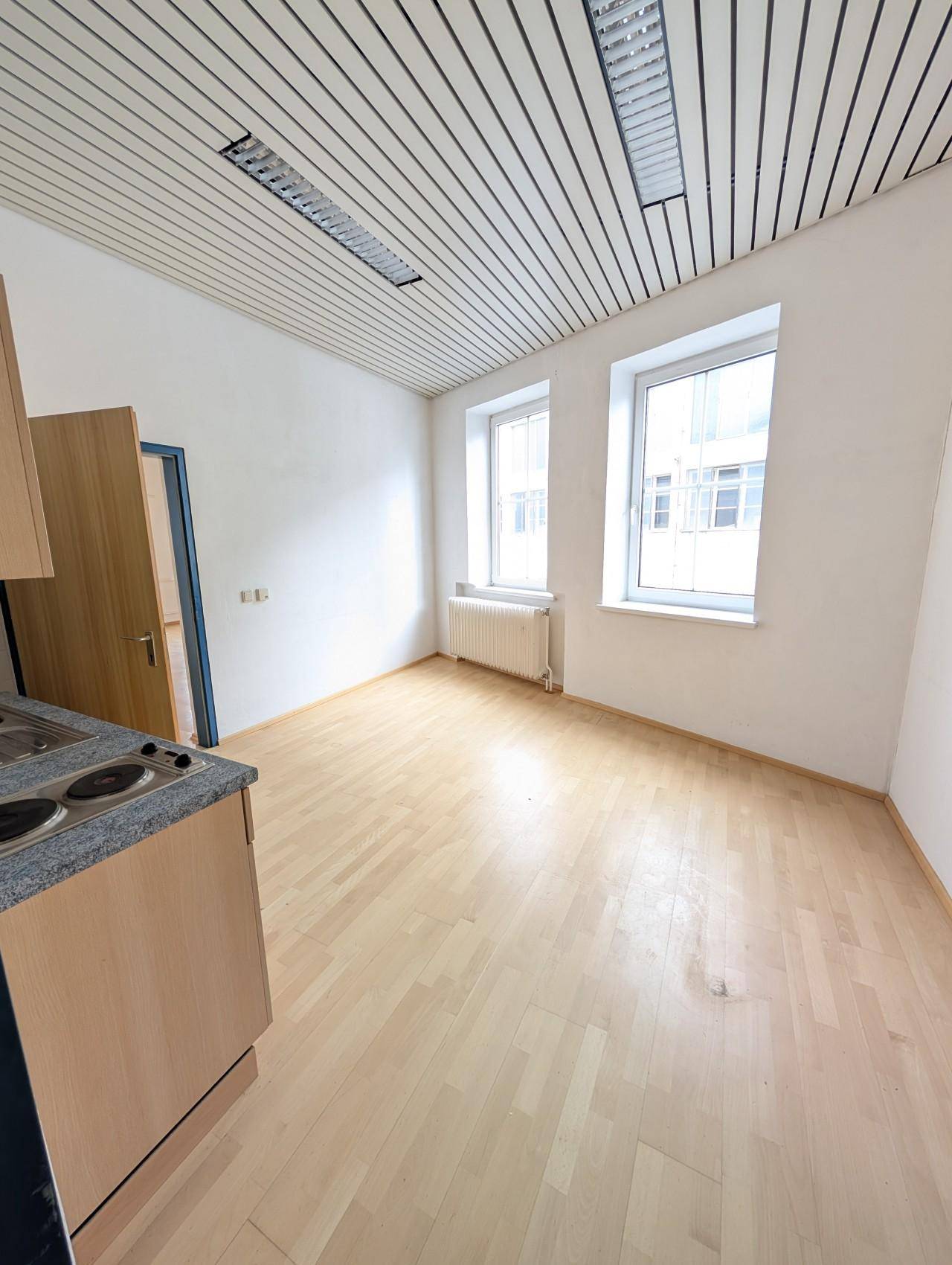 Kleiner Raum mit Holzboden, zwei Fenstern und einer einfachen Küchenzeile.