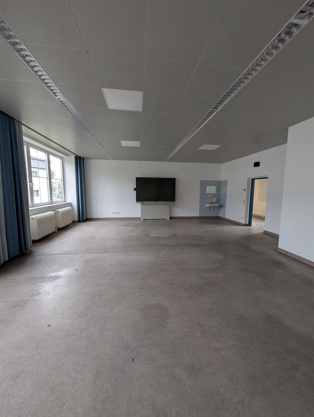Großer, leerer Raum mit Linoleumboden, abgehängter Decke und großen Fenstern.