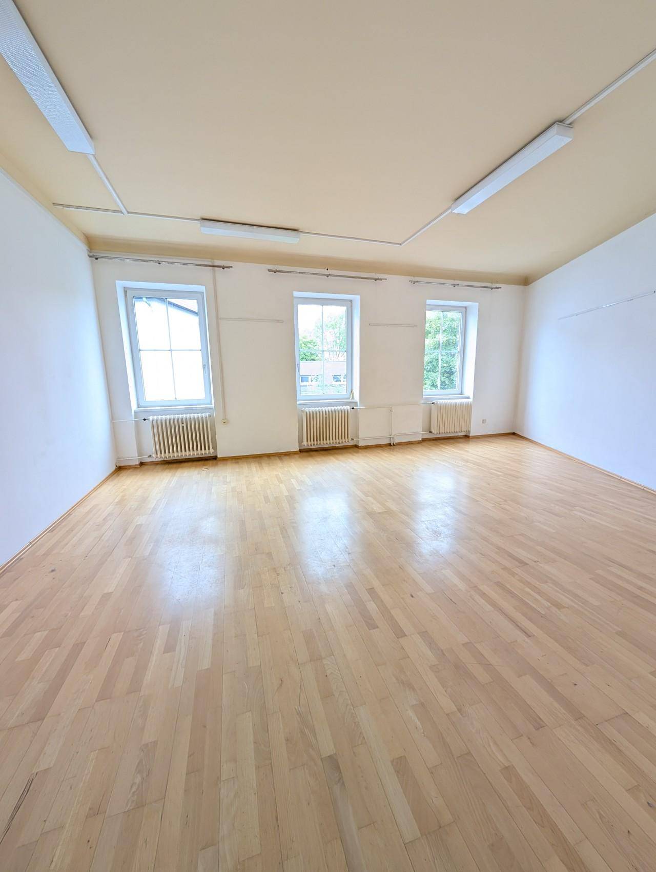 Heller, leerer Raum mit Holzboden, drei Fenstern und weißen Wänden.