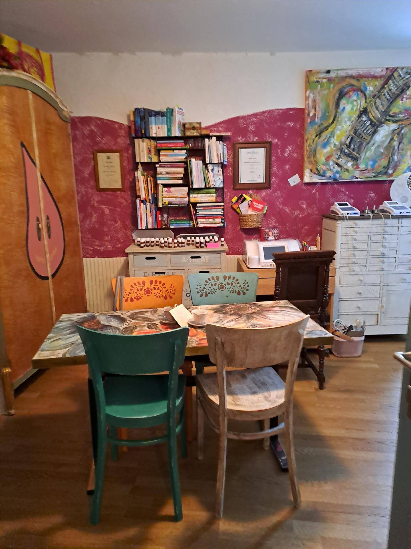 Essbereich mit einem Holztisch, bunten Stühlen, einem großen Schrank und Bücherregalen. Die Wand ist in Magenta gestrichen.