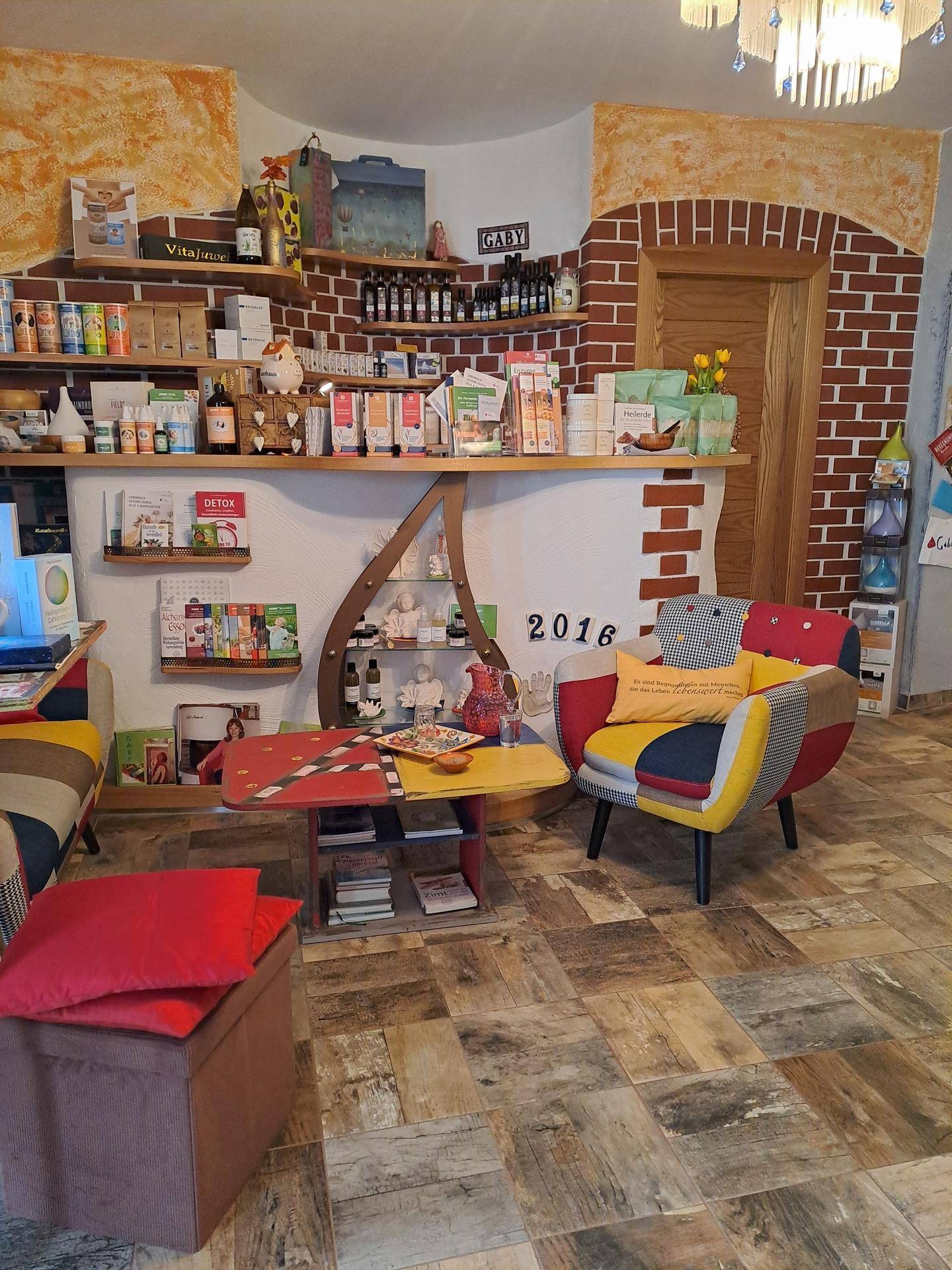 Heller Wohnbereich mit einem bunten Sessel, einem Couchtisch und Regalen mit verschiedenen Produkten. Eine Wand ist im Ziegelmuster gestaltet.