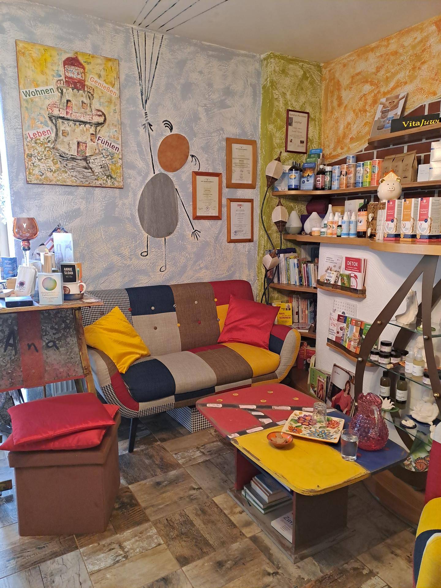 Gemütlicher Wohnbereich mit einem bunten Patchwork-Sofa, einem farbigen Couchtisch und Regalen voller Produkte. Die Wände sind künstlerisch gestaltet.