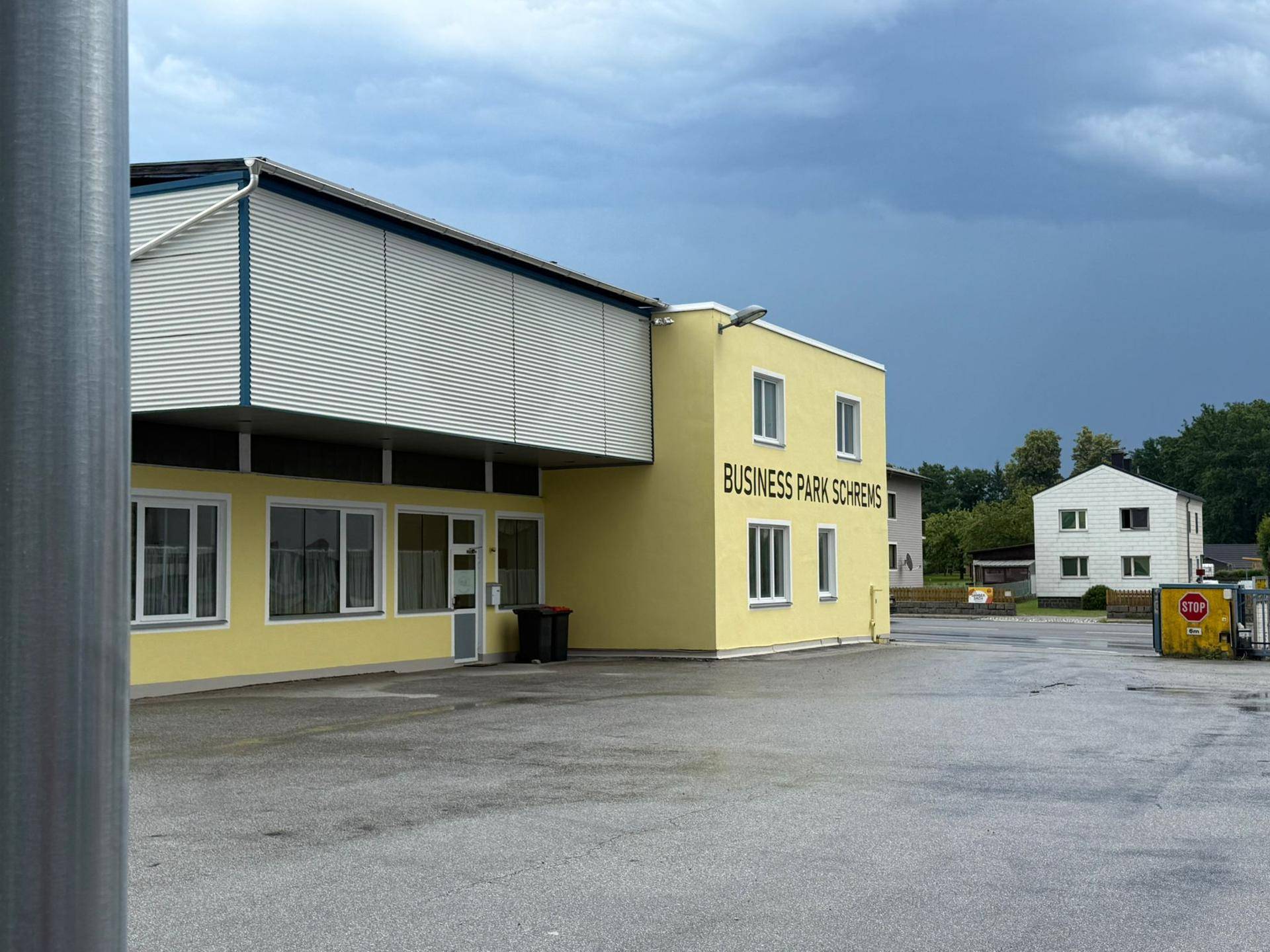 Gewerbegebäude mit gelber Fassade und Parkplatz, umgeben von weiterer Bebauung unter dunklem Himmel.