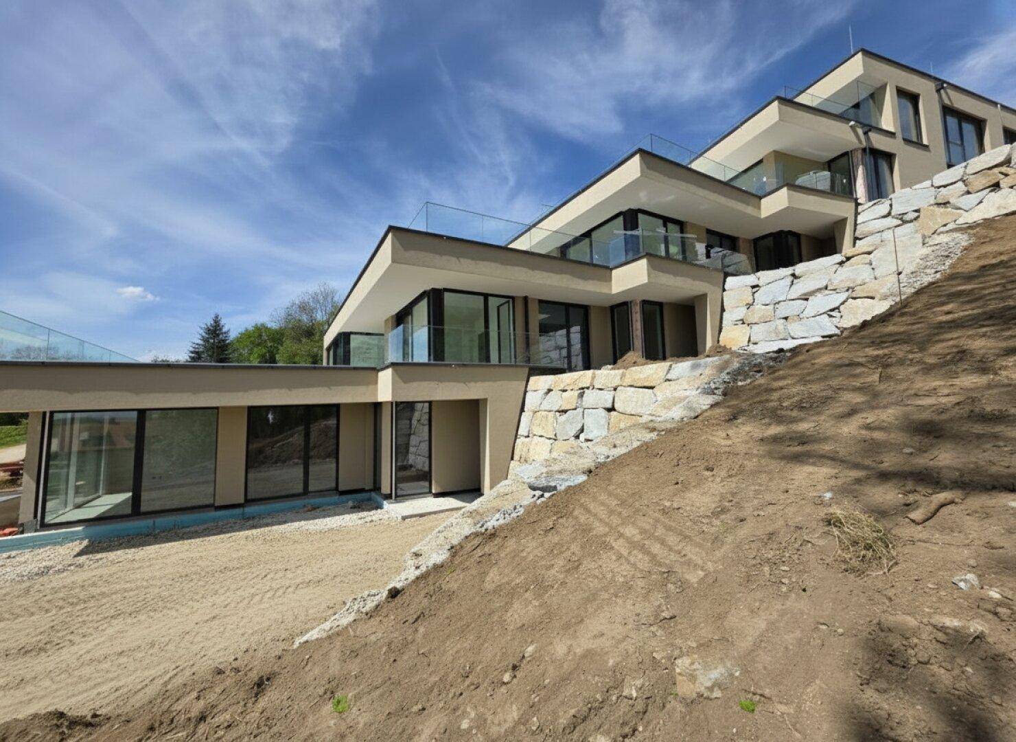 Terrassenhaus in Hanglage mit moderner Architektur, großen Glasfronten und Natursteinmauern im Bau.