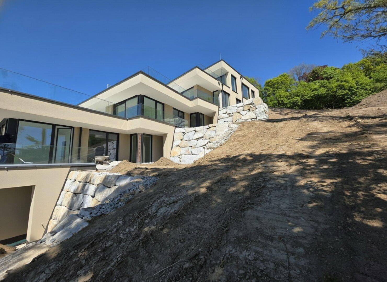 Modernes Terrassenhaus in Hanglage mit gestufter Architektur und Natursteinmauern unter blauem Himmel.