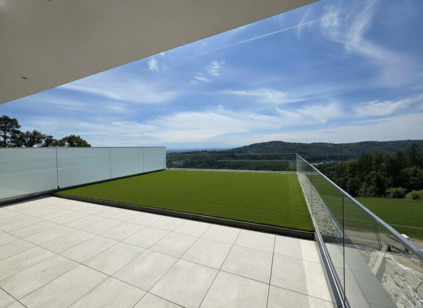 Moderne Terrasse mit Kunstrasenfläche und weitem Blick über die Landschaft unter blauem Himmel.