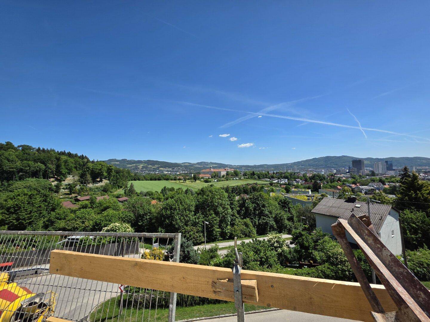 Panoramablick von der Baustelle über weite grüne Flächen, die Stadt und die umliegenden Hügel.
