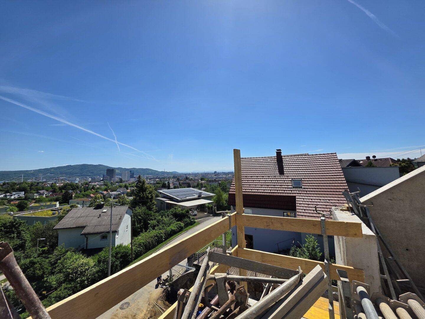 Weitblick von der Baustelle über Dächer, die Stadt und die hügelige Landschaft unter blauem Himmel.