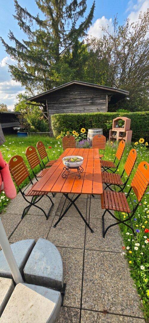 Gemütliche Terrasse mit Holztisch und Stühlen, umgeben von einem blühenden Garten und einem Gartenhaus.