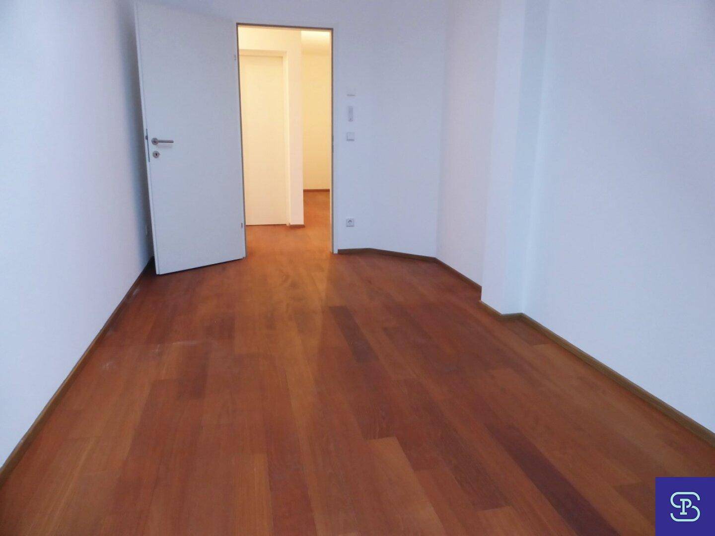 Geräumiges Zimmer mit Holzboden und weiß gestrichenen Wänden, Tür zum Flur.