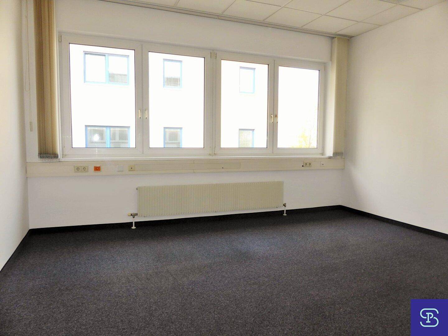Heller Büroraum mit großer Fensterfront, die den Blick auf ein gegenüberliegendes Gebäude freigibt.